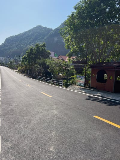 The image displays a serene roadside landscape in Trân Châu, Vietnam, captured on a clear, sunny day. A freshly paved asphalt road, featuring distinct yellow and white lane markings, stretches into the distance, occupying the right side of the frame. On the left, a majestic range of lush, green mountains dominates the horizon, rising under a bright blue sky.
Along the right side of the road, a line of dense green trees provides shade and separates the thoroughfare from the surrounding area. Nestled among these trees, various residential buildings with tiled roofs are partially visible. A small, rustic wooden structure, possibly a kiosk or shelter, stands close to the road. Adjacent to this structure is a sign with text in Vietnamese. The legible parts of the sign include "SƠN BÌNH ĐẶNG," "CÁP DUY TÂN - CÁP NHỰT TRANG," and a phone number (0936 900 472). Other smaller text is also present, likely detailing services or products. A metal railing or fence runs along the roadside beside the sign.
The absence of people or vehicles contributes to a sense of tranquility. The bright, clear lighting suggests it is either morning or early afternoon, with shadows indicating the sun's position. The scene evokes a feeling of peaceful travel through a scenic, mountainous region.