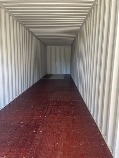 The image shows the interior of an empty shipping container. The walls and ceiling are ribbed and painted a light gray. The floor is made of dark red wooden planks with visible nail heads. The container is viewed from the front, looking towards the back. There are no people or any other objects visible inside the container. The scene appears to be located in Chakvi, Georgia, as indicated by the provided information.