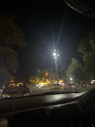The image depicts a view from inside a car at night, looking out through a rain-streaked windshield. The scene outside is illuminated by streetlights and the lights of businesses. Trees line the street on both sides. Several cars are visible on the road, some parked and some in motion. The overall impression is of a wet and dimly lit urban environment. The specific location of Batken, Kyrgyzstan, is not determinable from the visual information presented in the image.