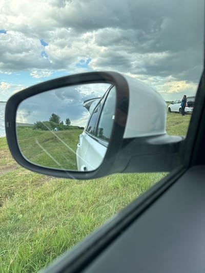 The image, captured in Айдарлы, Kazakhstan, is viewed from inside a vehicle, looking out the passenger side window and into the side-view mirror. The foreground consists of the dark interior trim of the car's door and window frame on the right and bottom, and a side-view mirror on the left and center.
Through the window, directly above the side-view mirror, a cloudy sky with patches of blue is visible. Further to the left, a body of water meets the horizon beneath the sky. To the right of the side-view mirror, a white car is parked on a grassy field, with its trunk open. A person, dressed in dark clothing and carrying a blue bag over their shoulder, stands facing the open trunk of this white car. The ground in the foreground outside the car is covered in green grass.
The side-view mirror reflects the scene directly behind the vehicle. This reflection shows a vast expanse of green grassy ground with faint tire tracks or paths. On the left side of the reflection, the body of water is visible. In the middle distance of the reflection, a cluster of small trees and bushes stands, with a light-colored, possibly blue, dome tent pitched nearby. The right side of the mirror's reflection shows the rear quarter panel and open trunk of the white car from which the photo is being taken, indicating it is parked near the water's edge.
