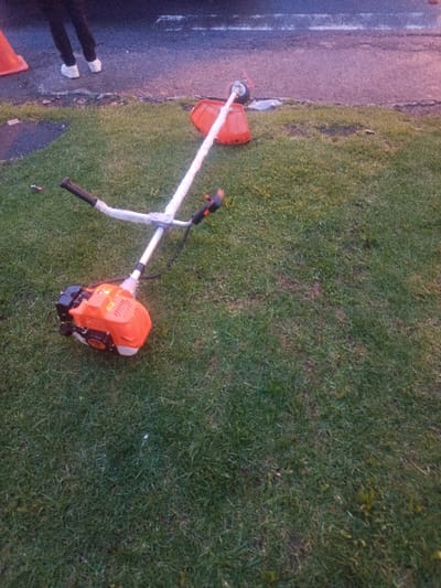 An orange and silver Stihl gas-powered string trimmer is depicted lying horizontally on a grassy area in Sangolquí, Ecuador. The trimmer's engine housing, fuel tank, and blade guard are orange, while the shaft is silver. Its handlebars are black. The device rests on green grass which exhibits some irregular brown patches. Immediately adjacent to the grass, a dark paved surface extends across the upper portion of the frame. On this paved surface, the lower legs and white-shoed feet of one individual are visible, standing near a partially visible orange traffic cone located on the far left. A faint white line marking is discernible on the paved surface, which reflects a purple and pinkish ambient light, suggesting a low-light environment such as dusk or dawn. No active use of the trimmer or interaction between the person and the objects is depicted.