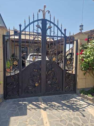 The image shows an ornate, black wrought-iron gate with gold accents, in front of a house in Nor Kharberd, Armenia. The gate features intricate scrollwork and pointed finials at the top. Behind the gate, a white car is partially visible, parked in a driveway or courtyard. The house has a tiled roof and beige stone walls. A rose bush with red flowers is in bloom to the right of the gate. The ground in front of the gate is paved with irregular grey stones, and shadows from the gate are cast across the paving. The sky is clear and blue.