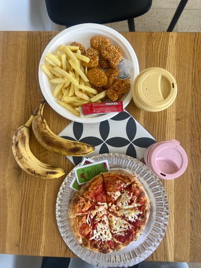 The image displays a wooden table laden with food and beverage items in Rosh Haayin, Israel. In the upper center, a white disposable plate contains golden-brown French fries and approximately ten heart-shaped, breaded food items coated in sesame seeds. A red ketchup packet and a small white sachet are also on this plate. To the right, a light yellow reusable cup with a matching lid is positioned. Below this arrangement, two ripe bananas, exhibiting significant brown spotting, rest directly on the wooden table surface. A black and white placemat featuring a geometric circle pattern is centered on the table. In the lower section, a small, personal-sized pizza, sliced into six pieces with a red sauce base and melted white shredded cheese, is presented on a silver-colored corrugated disposable plate. Adjacent to the pizza are two small green packets, labeled "CUP KETCHUP" in Hebrew and English. A pink reusable cup with a lid sits to the right of the pizza plate. The background in the upper left shows a white wall and a partial view of a black chair.