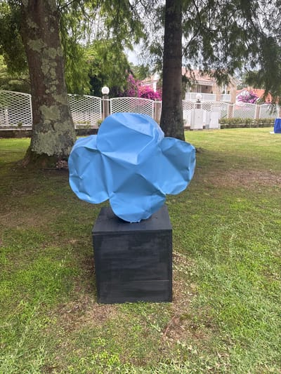 The image shows an outdoor scene in a grassy area, likely a park or garden. In the foreground, a bright blue, abstract sculpture with a crumpled, cloud-like appearance rests on top of a dark, rectangular pedestal. The sculpture is positioned slightly off-center to the left. Behind the sculpture and pedestal, a large tree with textured bark and green foliage dominates the left side of the frame. In the background, a white lattice fence separates the grassy area from a residential area with houses visible, featuring red tiled roofs and white railings. There are also vibrant pink flowers blooming along the fence line. Several light posts and electrical boxes are visible near the fence. The ground is covered in green grass with some patches of dry earth. The sky appears overcast. The overall impression is of a public outdoor space featuring a contemporary art installation, with a backdrop of residential architecture and natural elements. The location is specified as O Grove, Spain.