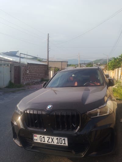 A black BMW SUV is parked on a paved surface in front of a brick wall and a metal gate. The car has orange interior seats visible through the windshield. The license plate reads "01 ZX 101 AM". Overhead power lines and utility poles are present in the background, along with houses and distant hills. The scene appears to be outdoors in a residential area, possibly in the village of Nor Geghi, Armenia. The lighting suggests it is daytime, with a slightly hazy sky.