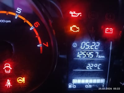 The image shows a close-up of a car's dashboard, illuminated by its own warning lights. On the left, a tachometer displays readings up to 6000 rpm, with the needle positioned between 5 and 6. Several illuminated warning lights are visible: a red oil can symbol, a yellow engine symbol, a red battery symbol, a red seatbelt symbol, a white symbol possibly indicating an open door, and a yellow steering wheel symbol with an exclamation mark. The digital display on the right shows the time as 05:22, a mileage of 125457 km, and a temperature of 22°C. Below this, a fuel gauge is partially visible. The bottom right corner of the image contains a timestamp: 25.07.2026 05:22. There are no people or environmental elements of the city of Mariinsky visible in this image.