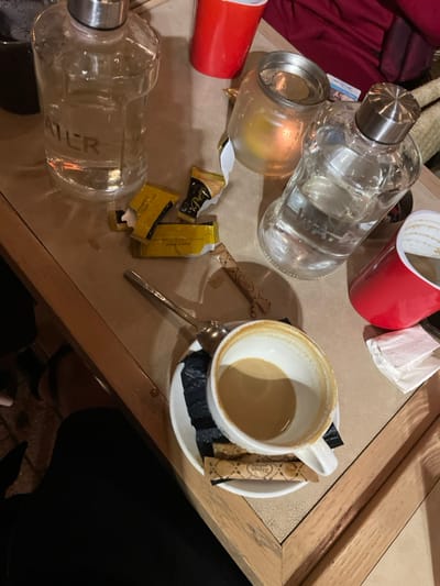 The image displays a casual tabletop scene, likely in an evening cafe or bar in Chaidari, Greece. The wooden table is seen from a top-down perspective, cluttered with various items suggesting recent use.
In the center of the frame, a white ceramic coffee cup holds a small amount of light brown liquid, presumably coffee with milk. It rests on a white saucer with a spoon and two brown sugar/sweetener packets, one showing partial text "Zarpan". Nearby, a small glass candle holder with a lit candle emits a warm, ambient glow, indicating a dim lighting environment, likely evening or night.
Two clear water bottles are visible on the table; one has "WATER" printed vertically on its side. There are also two red disposable cups, one upright and another partially visible, along with a discarded white napkin or tissue. Several yellow and gold packets, likely containing sugar or artificial sweetener, are scattered across the table; some are open, and one has "P...ice" visible on its packaging.
No faces are visible, but a leg is partially seen in the bottom right corner, suggesting someone is seated at the table. The collection of items points to a relaxed, informal gathering or a moment of leisure, perhaps after a coffee and drinks.
