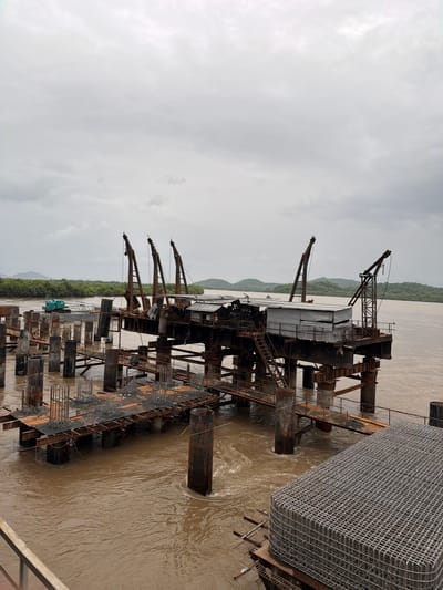 The image depicts a construction site in Vasai-Virar, India, likely on the Vasai Creek, a tidal estuary. A large, partially constructed bridge or pier structure dominates the scene, extending into the murky, brown water. The structure is supported by numerous large, rusty, cylindrical pillars. On top of the central platform of the structure, there are several industrial cranes, some appearing old and weathered, with visible cables and mechanical parts. A corrugated metal shed or building is also present on the platform.
In the foreground, to the right, is a large, rectangular cage-like structure filled with metal rebar, suggesting ongoing construction. To the left, more support pillars and scaffolding are visible, with metal rods protruding from concrete bases. In the middle ground, on the water, a turquoise construction vehicle with an extended boom, possibly a crane or excavator, is positioned on a barge or pontoon, actively engaged in work.
The background shows a lush, green, tree-covered coastline with hills in the distance under a heavily overcast, grey sky. The water is choppy, indicating a tidal current or wind. No people are clearly visible in the frame, but the presence of construction equipment and materials strongly implies human activity related to the project. The overall impression is one of an active, large-scale infrastructure project in progress, surrounded by a natural, watery environment.