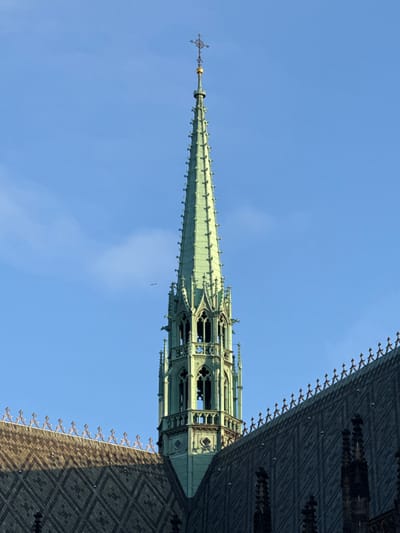 The image displays the upper section of the main south tower spire of St. Vitus Cathedral in Praha 1, Czechia, set against a clear blue sky with minimal cloud cover. The spire exhibits a verdigris green patina, indicative of oxidized copper or bronze, and tapers sharply upwards. It is crowned by a golden orb supporting an elaborate, dark metal cross finial. The spire's structure is detailed with numerous crockets along its edges and features an openwork belfry section below, characterized by pointed arches with tracery, within which dark bell-like objects are partially visible. Gargoyle figures protrude from the belfry's corners. Portions of two steeply pitched roofs belonging to the cathedral are present in the lower frame, covered with dark grey tiles arranged in a geometric diamond pattern. Decorative finials adorn the ridgelines of these roofs. The left roof section is partially illuminated by direct sunlight, while the right section is predominantly in shadow. A small, dark, unidentified airborne object is observed in the sky to the left of the spire. No people, discernible actions, or interactions are visible.