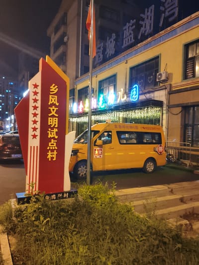 The image depicts a nighttime scene in Jiguan District, China. A prominent red and white sign with yellow text stands in the foreground, displaying Chinese characters that translate to "Xiangfeng Civilized Model Village." To the right of the sign, a yellow school bus is parked. The bus has Chinese characters and a phone number printed on its side. A red stop sign is visible on the rear passenger door. In the background, a building with yellow walls features a large blue neon sign with Chinese characters that appear to read "Guancheng Blue Lake." The building is illuminated by strings of small lights. A red flag on a flagpole is also visible. The environment appears to be a street or public area, with grass and vegetation in the foreground and a street or sidewalk behind the sign. The overall impression is that of a well-lit, urban environment at night.