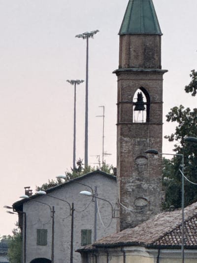 The image depicts a street scene in Fontevivo, Italy, featuring a prominent bell tower with a green, pitched roof. The tower, constructed of aged brick, has an arched opening revealing a bell. To the left of the tower, a light-colored building with a slanted roof and two windows is visible. Multiple street lamps with curved arms are positioned along the street. In the background, two tall utility poles with multiple lights are present. The sky is overcast. The scene appears to be a daytime shot, with no people or animals visible. The overall impression is one of a quiet, possibly historical, urban or semi-urban environment.