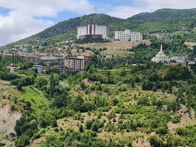 The image shows a hillside overlooking the city of Artvin, Turkey. The hillside is covered with lush green trees and vegetation, with several buildings scattered across it. In the background, a large white building with many windows stands prominently on the top of the hill, accompanied by a smaller white building. Closer to the foreground, a distinctive white mosque with a golden dome and minaret is visible. Several other residential and institutional buildings are also present on the slope. The sky is partially cloudy, with patches of blue sky visible. The overall scene depicts a settlement nestled within a verdant natural landscape.