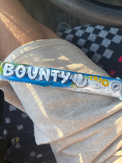 A Bounty Trio chocolate bar with three bars inside is held in the lap of a person wearing light-colored shorts. The chocolate bar is blue and white with the "BOUNTY" logo in large white letters and a depiction of a coconut. The text on the wrapper includes "TRIO 3X" and a Rainforest Alliance seal. The background shows a person's leg and the interior of a vehicle, with a checkered pattern visible. The scene appears to be outdoors during the day with sunlight casting shadows. There is no visible indication of the city of Zlataritsa, Bulgaria in the image.