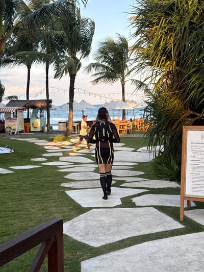 Here is a detailed description of the image:
The image is an outdoor scene at Hotel Komune Beach in Medahan, Indonesia. The setting appears to be a beachside resort or club. Palm trees are prominently featured in the foreground and background. The sky is visible, suggesting daytime.
A person in a black and white wetsuit with the word "USWE" visible is standing on a paved pathway that is white with green patches. Another person is standing to their side. The pathway has a pattern of irregular-shaped stones or concrete pieces.
In the background, towards the ocean, there are several white parasols and wooden chairs set up, indicating a relaxation area. A small food or beverage kiosk is also visible.
To the side of the image, there is a wooden sign displaying information, seemingly regarding hotel amenities or rules.