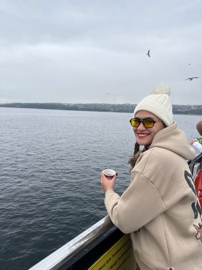 An adult female is depicted on a vessel, likely a ferry, navigating a body of water near Darıca, Turkey, possibly the Marmara Sea or Izmit Bay, under an overcast sky. She is positioned on the right side of the frame, smiling and looking towards the camera. She wears a white knitted beanie with a pompom, yellow-tinted aviator-style sunglasses, and a beige hooded sweatshirt with a dark graphic design partially visible on the back. Her dark hair is styled in plaits over her left shoulder. In her right hand, she holds a small, dark-colored disposable cup. She leans against a metallic railing that extends horizontally across the foreground. The water behind her is dark and shows ripples. A distant coastline with low-rise structures is visible on the horizon. Several seagulls are scattered across the cloudy sky. A yellow sign with partially visible Turkish text is present below the railing. Partial view of another individual's green and white clothing is visible on the far right.