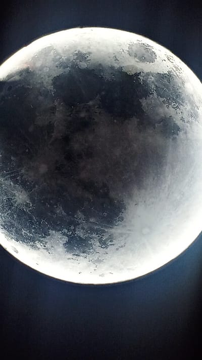 The image is a close-up, vertical shot of the moon against a dark background. The moon is partially illuminated, with a bright, white crescent shape on the right side, gradually fading into shadowed areas on the left. The surface of the moon is clearly visible, showing craters, textures, and variations in light and shadow. The dark background suggests the moon is viewed at night, likely in an outdoor setting, devoid of any city lights or structures that would indicate it's in Choktal, Kyrgyzstan. There are no people, subjects, objects, or actions depicted in the image.