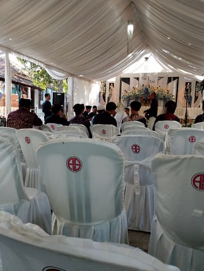 The image displays a daytime event taking place under a large white fabric tent structure in Juwana, Indonesia. Approximately twenty individuals are visible, primarily men, seated in white-covered chairs arranged in multiple rows facing a decorated stage. Many chair covers feature a circular red logo containing the letters "AS." The tent ceiling is draped with white fabric, from which a slender, ornate light fixture hangs centrally.
The stage area at the far end is adorned with white panels, blue and white floral arrangements, and vertical crescent moon-shaped light fixtures. A table is positioned on the stage. Several individuals are standing on or near the stage, including one man in a dark shirt and another in a lighter shirt. One individual at the front, partially obscured by other attendees, wears a white head covering.
On the left side of the tent, a man in a dark shirt stands near a black speaker unit. Beyond the tent structure on the left, traditional residential buildings with tiled roofs, wooden walls, and light blue window or trim details are visible under clear skies. The individuals in the foreground are observing the proceedings on the stage.