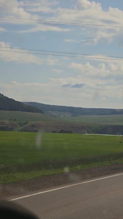 The image depicts a rural landscape under a partly cloudy sky, likely in or near the village of Kama-Ismagilovo, Russia, given the context. The foreground features a paved road with a white stripe. Beyond the road, a green field stretches towards rolling hills. In the middle ground, several small structures, appearing to be houses or farm buildings, are visible scattered across the landscape. The background is dominated by a dense forest covering a hill. Overhead, two thin power lines cross the frame. The overall scene is tranquil and natural, characteristic of the Russian countryside.