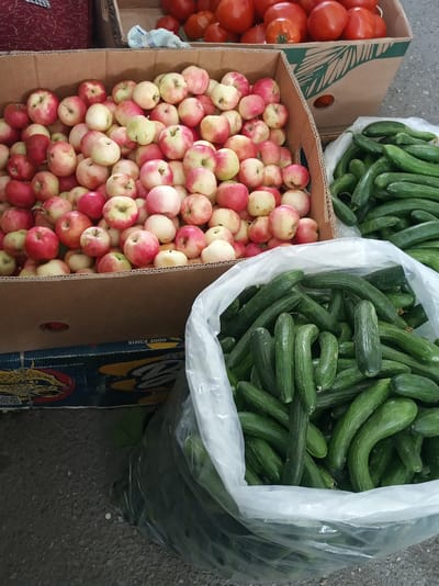 In this image, a market stall displays a variety of fresh produce. The foreground is dominated by a large clear plastic bag filled with numerous green cucumbers. To the left of the cucumbers, a cardboard box overflows with a colorful assortment of apples, predominantly red and yellow. In the background, another cardboard box is visible, containing bright red tomatoes. The setting appears to be an outdoor market, with a rough, grey paved ground. The overall impression is one of a bountiful harvest and a bustling marketplace. There is visible text on the lower left side of the image: "SINCE 2000". The lighting suggests it is daytime.