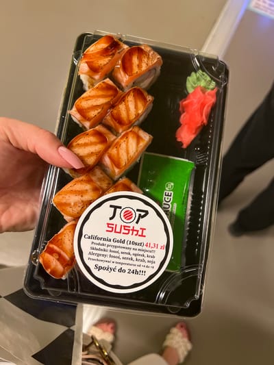This media file captures a close-up view of a person's hand holding a plastic container of sushi. The sushi appears to be California rolls, topped with grilled salmon and a glaze. The container also holds small portions of pickled ginger and wasabi. A circular label on the container provides product information, including the name "California Gold," the quantity "10szt" (10 pieces), and the price "41,31 zł" (41.31 Polish Zloty). It also lists ingredients and allergens in Polish, such as "łosoś" (salmon), "serek" (cheese spread), "ogórek" (cucumber), "krab" (crab), and "soja" (soy). Instructions for storage and consumption are also present: "Przechowywać w temperaturze od +4 do +8" (Store at temperatures from +4 to +8) and "Spożyć do 24h!!!" (Consume within 24 hours!!!). A small packet of soy sauce is visible in the background. The scene is likely indoors, given the lighting and the presence of a person wearing slippers. The background suggests a casual setting, possibly a home or a takeaway environment. The overall impression is one of a ready-to-eat meal.