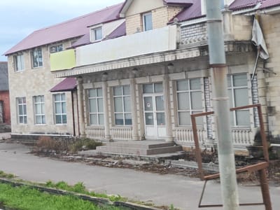 The image depicts the exterior of a weathered two-story building in Zavidovo, Russia. The building features a prominent red corrugated metal roof and walls constructed from light-colored stone. A large, faded banner is stretched across the upper facade, partially obscured by a street lamp pole in the foreground. The lower level of the building has a row of large windows and a central entrance with steps, all decorated with ornate white balustrades and columns. Surrounding the building, the environment appears neglected, with debris scattered on the ground and sparse vegetation. A rustic metal frame stands on the pavement in front of the building, and a tattered white flag hangs from the right side of the facade. The overall scene suggests a commercial or administrative building that is either disused or in a state of disrepair.