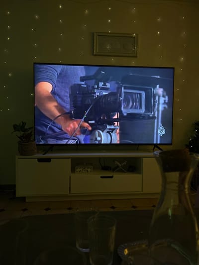 A television screen displays a person holding a professional video camera. The screen is mounted on a wall adorned with string lights and a framed artwork. To the right of the screen is a white cabinet with decor items inside. A wine bottle and glasses are visible, out of focus, reflecting on a dark surface. A small potted plant is placed near the bottom of the cabinet.