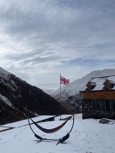This image captures a snowy mountain landscape in Juta, Georgia, under an overcast sky. In the foreground, a multi-colored fabric hammock, primarily blue, red, and yellow, rests on a dark, curved freestanding stand, with fresh snow partially covering the hammock and the ground around it. Behind the hammock, positioned centrally, a white Georgian flag with a large red cross and four smaller red crosses in its quadrants flies from a dark flagpole, rippling slightly. To the right of the flag, a two-story building with a stone base and a wooden upper section is visible. Its roof is covered in snow and features two gabled dormer windows. The building has multiple large rectangular windows. The background is dominated by snow-covered mountains. A dark, rocky and sparsely vegetated slope, partially dusted with snow, rises on the left, contrasting with a heavily snow-covered peak on the right. Additional smaller, snow-dusted structures with simple roofs are discernible in the mid-ground to the left of the flagpole. The entire ground plane is covered in a layer of white snow, showing some tracks.