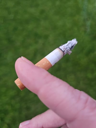 The media file shows a close-up of a hand holding a lit cigarette. The cigarette is partially burnt, with ash at the tip. The hand is fair-skinned and appears to be a person's index finger and thumb holding the cigarette. The background is a blurred green surface, likely grass, indicating an outdoor setting. The lighting suggests it is daytime, but no specific weather conditions are apparent. The image is focused on the cigarette and the fingers, with the background out of focus. There is no visible text in the image. The context provided indicates the location as Mäntsälä, Finland. The activity taking place is someone holding a lit cigarette.
