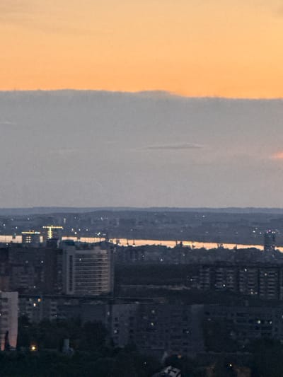The image shows a twilight cityscape, likely Shirkovo, Russia, as indicated. The sky is a gradient of orange and grey, suggesting either sunrise or sunset. A prominent, thick grey cloud band stretches horizontally across the middle of the frame. Below this, a darker band of land with subtle lights is visible, possibly a distant cityscape or industrial area. The foreground is dominated by buildings, appearing to be apartment complexes. They are dark and dense, with some windows illuminated, suggesting occupancy. The overall impression is a quiet, urban scene at dusk or dawn.