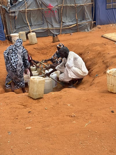In Umm Sudayra, Sudan, individuals are gathered around a communal water source. The scene depicts a sandy, outdoor environment with several large, plastic water containers. Tents, likely temporary shelters, are visible in the background. People are dressed in traditional clothing and appear to be collecting water from spigots attached to pipes. One person is crouched near the water containers, while others stand and tend to the filling of their containers. The overall impression is one of a community engaged in a daily necessity, likely in a place experiencing resource scarcity.