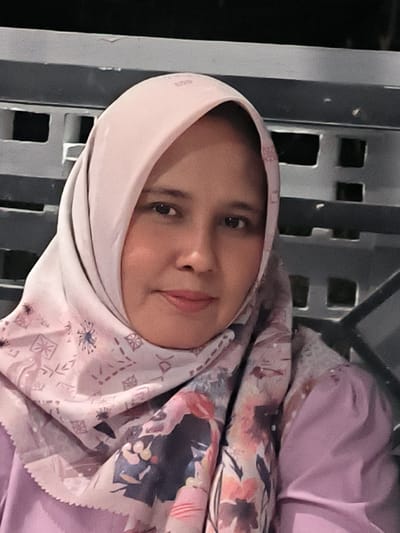 A medium shot, waist-up photograph captures a woman wearing a light pink hijab and a lavender top, with a patterned scarf draped over her shoulders. The scarf has a floral design in shades of pink, blue, and black, with a border of small, repeating pink geometric shapes. The woman has medium-toned skin, dark eyes, and a slight smile. She is positioned in front of a dark, metal, grid-like structure, possibly a fence or railing, that is out of focus. The background is dimly lit, suggesting an evening or indoor setting. There are no discernible landmarks or environmental cues to pinpoint a specific location within Panteraya, Indonesia. The overall lighting is soft, and the focus is primarily on the woman's face and upper body.