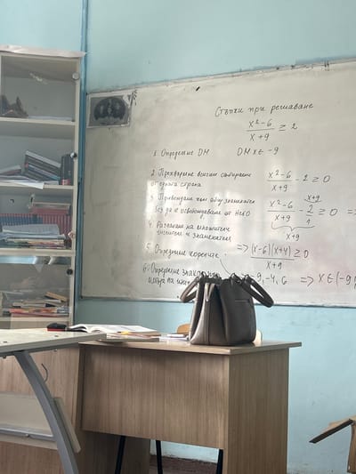 The scene depicts a classroom in Dolni Tsibar, Bulgaria. A white board on a light blue wall dominates the center, displaying mathematical equations and steps written in Bulgarian script, specifically concerning the solution of the inequality `(x^2 - 6) / (x + 9) >= 2`. To the left of the white board, a multi-shelved, light-colored bookcase is visible, filled with numerous books, binders, and papers. Above the top left corner of the white board, a small, framed black and white photograph depicting three or four individuals is affixed to the wall. In the foreground, a light brown wooden desk occupies the right side, upon which a grey or taupe-colored handbag with two handles is placed. To the immediate left of this desk, the corner of another desk with a light-colored top and a grey metal frame is partially visible, featuring an open book. No individuals are present in the image.