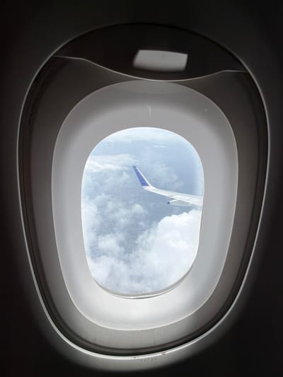 The image shows a view through an airplane window. The window has a distinctive oval shape with layered framing, typical of aircraft cabins. Through the window, we can see a vast expanse of sky filled with white, fluffy clouds. A portion of an airplane wing, with blue and white markings, is visible extending from the right side of the frame into the cloudy sky. The lighting suggests it is daytime, likely mid-flight. There are no people or specific city landmarks visible in this image. Therefore, it is not possible to determine a location within the city of Bhiwandi, India, based solely on this photograph.