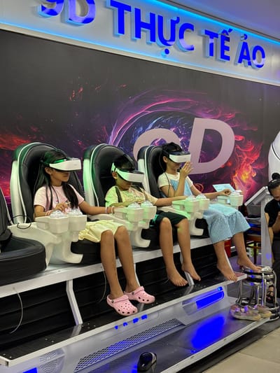 Three young children are seated in a row of futuristic-looking chairs, each wearing a virtual reality headset and appearing engrossed in an immersive experience. The chairs are white with black accents and have integrated control panels. The backdrop features a large, abstract graphic with swirling colors and a prominent "3D THỰC TẾ ẢO" (3D Virtual Reality) sign in blue, illuminated lettering. The scene suggests an entertainment venue, possibly an arcade or amusement center, located in Phu Thuy, Vietnam. The children are dressed casually, and their feet are visible, with one child wearing pink clogs. To the right, another person is partially visible, looking down at a device. The lighting is ambient with blue lights illuminating the lower sections of the seating area.