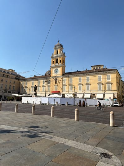 The image captures a sunny day in a European city square, likely Parma, Italy. The main subject is a tall, yellow building with a clock tower. The building features numerous windows with closed shutters and white awnings. A man is seen cycling past the building on a cobblestone street. Several white tents are set up in front of the building, possibly for a market or event. Stone bollards are placed along the edge of the square, and a shadow of a street lamp is cast across the paving stones. The sky is a clear, bright blue, suggesting good weather. A crane is visible in the background, indicating construction or renovation work. The scene appears to be a public gathering space in a historic urban setting.