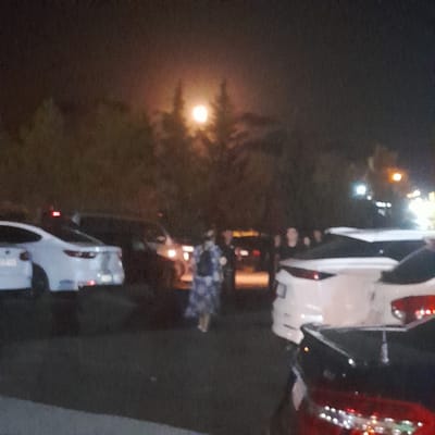 The image depicts a nighttime scene in what appears to be a parking lot. The primary subject is a full moon visible through silhouetted trees in the upper center of the frame, casting a bright, diffuse light. In the foreground, several vehicles are parked, predominantly white and dark-colored SUVs and sedans, some with their brake lights illuminated. A person wearing a patterned garment, possibly a dress or robe, is walking away from the viewer towards the right, positioned between several vehicles. The environment suggests an outdoor, possibly semi-rural or suburban setting, given the presence of trees and a parking area. The overall lighting is dim, with the moonlight and vehicle lights being the main sources of illumination, creating a somewhat grainy and low-contrast image. The location in Rudəkənar, Azerbaijan is noted, though the visual evidence within the image itself does not confirm or deny this specific location.