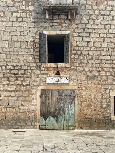 This is a medium shot, eye-level, exterior, static, daylight photograph taken in the city of Grad Stari Grad, Croatia. The image depicts the stone facade of a building. In the center of the facade, there is a wooden door with peeling green and brown paint, and two metal hinges. Above the door, a white sign with the words "LAJANIS HOME design" and a depiction of a rooster is mounted. Centered above the sign is a small, rusted bell hanging from a bracket. Above the bell, an open window with dark shutters shows a dark interior. The building is constructed of large, irregular, light-colored stones with visible mortar. The ground in front of the building is paved with stone slabs. A dark metal grate is visible on the ground near the bottom left of the image.