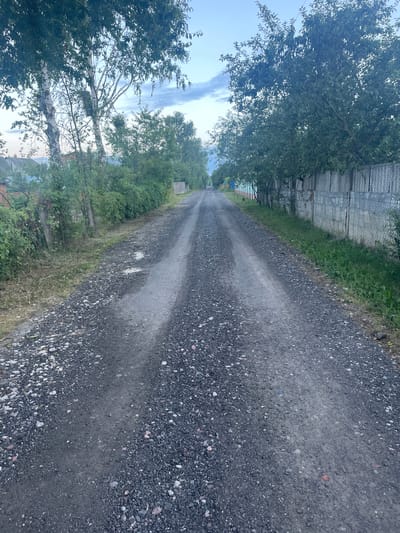 An outdoor, low-angle, vertical, full shot shows a gravel road in Babovitski Rural Council, Belarus. The road is centered and leads into the distance, with a slight curve to the left. The ground is covered in dark gray gravel and dirt, with some lighter patches and puddles of water visible. Trees with green foliage line both sides of the road. On the left, a wooden fence and some bushes are visible, with houses in the background. On the right, a wooden fence made of planks and wire mesh runs along the edge of the road, with more trees and bushes behind it. The sky is a pale blue with some wispy white clouds. The overall scene depicts a rural road in what appears to be a village or settlement.