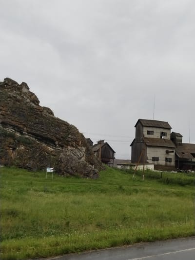 The image depicts a rural scene in the city of Lakly, Russia, featuring a rocky outcrop on the left and industrial buildings on the right. The rocky formation is layered and appears to be a natural geological feature. Adjacent to the rock, a small sign is visible on a post, likely providing information about the area.
In the middle ground, several rustic wooden and corrugated metal buildings stand. One prominent structure is a multi-story building with a sloping roof, possibly a grain elevator or an industrial facility. Another smaller wooden building is situated to the left of this main structure. The buildings appear weathered, suggesting age and exposure to the elements.
The foreground consists of a lush green grassy field, dotted with wildflowers, sloping gently towards the viewer. A paved road with a white line is visible at the bottom of the image, indicating its presence near a thoroughfare. The sky is overcast with a uniform gray cloud cover, suggesting a dull or cloudy day. There are no people visible in the image. The overall impression is one of a functional, somewhat isolated industrial area situated within a natural landscape.