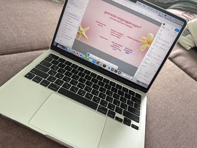 The image shows a close-up of an open silver Apple MacBook laptop resting on a textured, rose-colored fabric surface, likely a couch. The laptop screen displays a design software interface, possibly Figma, with a pink background and Russian text elements. Text boxes on the screen contain phrases like "для кого подходит курс?" (who is the course for?), "наш курс идеально подойдет:" (our course is ideally suited for:), and lists of target audiences. A starfish and a yellow flower are superimposed on the design as visual elements.
On the left side of the screen, a layers panel is visible, showing various design elements. The keyboard of the laptop is clearly visible, with Cyrillic and Latin characters on the keys. The trackpad is also visible. The overall lighting suggests an indoor setting, possibly a home environment.
While the image is a close-up of a laptop, there are no indications or elements within the image that place it specifically in the city of Коробицыно, Russia. The description is purely based on the visual content presented.
