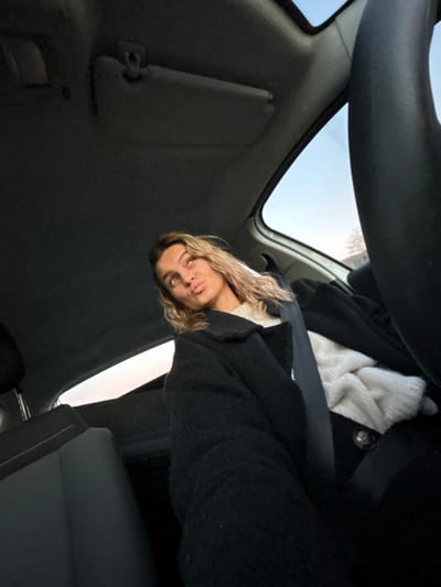 A woman in a car wears a black coat over a white sweater. She's wearing a seatbelt. The view looks up towards the sunroof, which shows a glimpse of the sky. A steering wheel is at the top of the frame. The car's interior is visible, including the headliner and part of a sun visor.