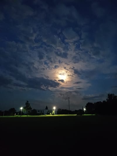 The image captures a nighttime scene in Trypillia, Ukraine, featuring a full moon partially obscured by clouds. The moon casts a soft glow on the surrounding environment, illuminating a dark, grassy field in the foreground. In the mid-ground, a low-rise building with lit windows and several streetlights can be seen, casting pools of light onto the field. Trees and a utility pole are also visible. The sky is a deep blue, with scattered clouds creating a dramatic contrast with the bright moon. The overall atmosphere is serene and tranquil.