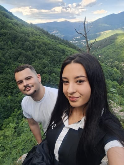 A selfie photograph of a young adult woman and man is taken in an outdoor setting, with a scenic mountain backdrop.
The woman, positioned in the foreground at the top of the frame, has long, dark hair and is wearing a black and white top. She is slightly smiling towards the camera.
The man, positioned in the lower portion of the frame, is wearing a white t-shirt and is looking towards the camera with a slight smile. He has a mustache and short, dark hair.
Behind the couple, a dense forest is visible, transitioning into distant mountain ranges under a partly cloudy sky. The image is taken from a high vantage point, possibly near or in the city of Zasele, Bulgaria. The picture is rotated clockwise.