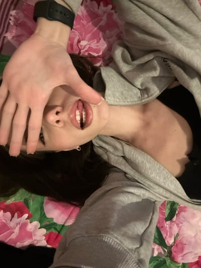 The image displays a young female, likely indoors within a residential setting in Волховец, Russia. She is observed from an overhead perspective, lying on her back. Her left hand is raised, palm facing the camera, with fingers outstretched to partially obscure her face, specifically her eyes and forehead. A dark-colored smartwatch is present on her left wrist. Her lips are parted, revealing both upper and lower teeth, with the tongue partially visible. She has dark hair, which is spread out on the surface beneath her head. She is dressed in a light gray hooded sweatshirt, partially unzipped, exposing a dark garment beneath. The background consists of a textile surface patterned with prominent pink floral designs, resembling a bedspread or blanket.