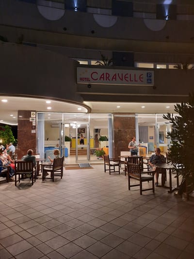 The image shows the entrance and outdoor seating area of the Hotel Caravelle in Pesaro, Italy. The hotel's sign is visible above the entrance, featuring the name "CARAVELLE" in red lettering with a parking symbol. The entrance itself is a large glass door with a reception area visible inside, where a person in dark clothing stands behind a counter. Several people are seated at tables on the tiled outdoor patio in front of the hotel. Some are engaged in conversation, while others appear to be relaxing. The area is lit by overhead lights, and the overall atmosphere suggests an evening setting. The architecture of the hotel features curved balconies and modern elements.