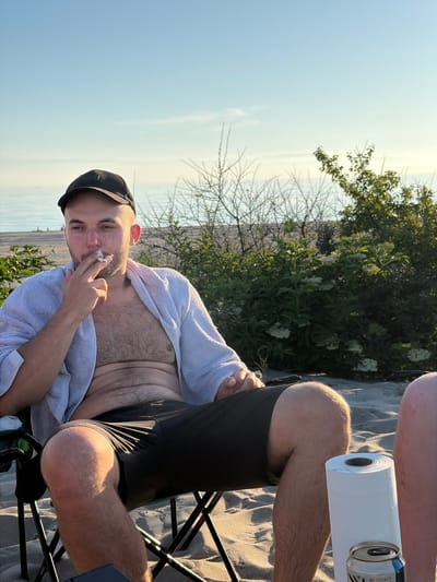 A man wearing a black baseball cap and a white towel draped over his shoulders sits in a folding chair on a sandy beach. He is smoking a cigarette and looking to his left. He is wearing dark shorts. In front of him is a roll of paper towels and a can. Behind him are some bushes and a body of water. The sky is clear and blue, with a faint hint of clouds. The location is identified as Povarovka, Russia.