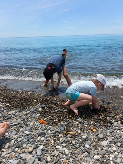 The image depicts a sunny pebble beach scene in Krinitsa, Russia. In the immediate foreground, a bare human foot rests on light-colored, smooth pebbles. To the right, a young child, wearing a light blue short-sleeved shirt, blue and green patterned swim trunks, and a white mesh baseball cap, crouches barefoot at the water's edge, intently focused on the wet pebbles. Two small toy cars (one orange, one blue) and an orange boat-like toy are visible on the pebbles near this child. Slightly behind and to the left, an older child or pre-teen, wearing a dark blue short-sleeved shirt and a black baseball cap with a red "H" logo, bends forward with hands submerged in the shallow breaking waves, appearing to collect or examine items. Further into the calm, clear blue sea, two additional individuals are partially visible, with heads and shoulders above the water. The horizon line is clearly defined, separating the serene sea from a light blue sky with faint, wispy clouds. Small, white-crested waves break gently onto the pebble-covered shore.
