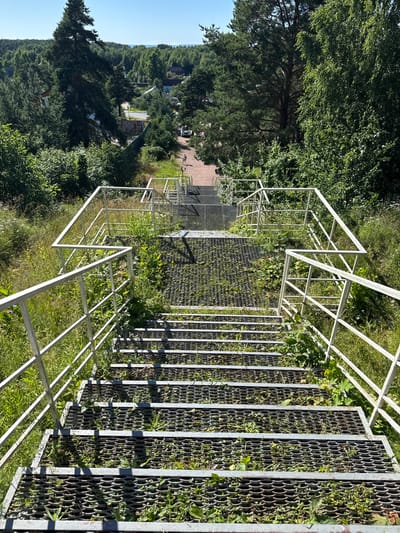 This is a daytime, high-angle, eye-level shot of a long, metal outdoor staircase in St. Ozerki, Russia, leading down a grassy slope. The staircase has a zigzag configuration with white metal railings on both sides. The steps are made of a perforated metal grate, and grass and weeds are growing up through them. The surrounding environment is lush and green, with trees and foliage visible on either side of the stairs. In the distance, at the bottom of the slope, there are some buildings and a road where two people are walking. A white car is parked on the side of the road. The sky is clear and blue.
