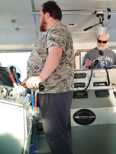 The photo depicts two men on what appears to be the bridge of a vessel. The first man, who is closest to the camera, has a beard and is wearing a t-shirt with writing on it and dark trousers. He is standing by a control panel. The second man is wearing sunglasses and a gray shirt with "Scotland" printed on it, is standing behind the control panel. Both men are indoors, and the light is fairly bright. There are various gauges, knobs, and other nautical equipment on the dashboard. There is no information to conclude that the image was taken in the city of Pavlokovo, Russia.