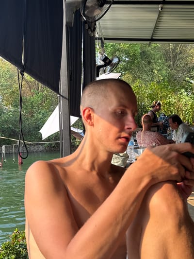 The image shows a shirtless man with a shaved head getting a haircut outdoors. The location appears to be a covered patio or deck with a view of a body of water and trees. Other people are present in the background at tables. Electric fans are visible on the deck. Cables and possibly a dock are visible along the water's edge. The image was taken during daylight. This location is in the city of Tambon Nam Phrae, Thailand.